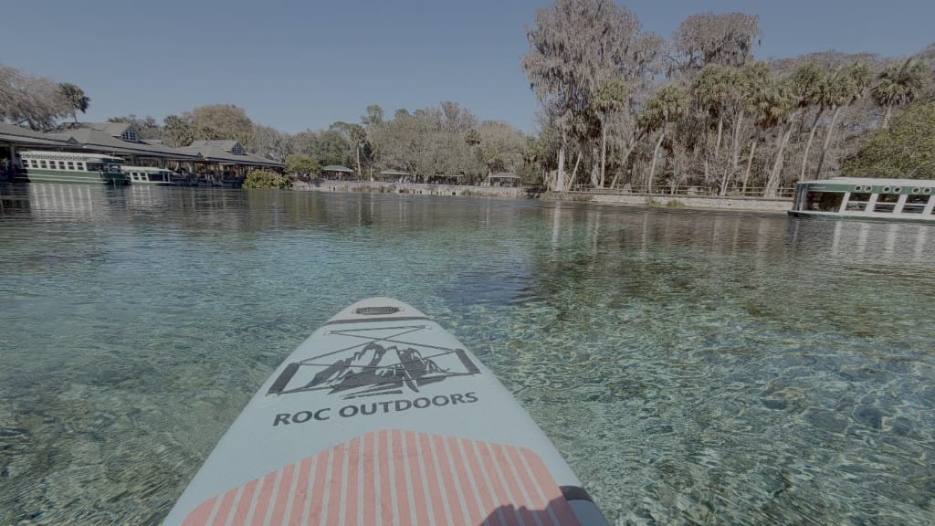 View from the nose of a ROC Outdoors paddleboard over clear spring water, with glass-bottom boats and trees along the bank
