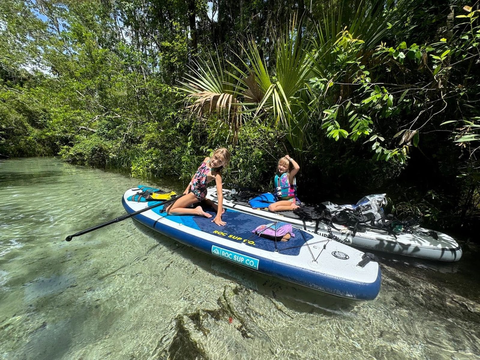 Two children in life jackets resting on a ROC SUP Co. paddleboard at a shaded spring bank