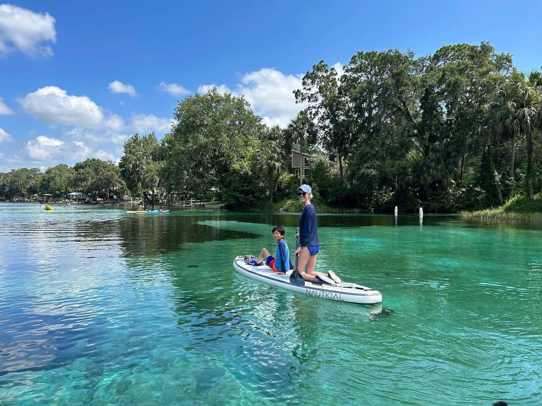 A guest standing on a paddleboard with a child sitting on the nose, on bright turquoise spring water
